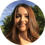 A woman with long brown hair smiles outdoors with trees and a blue sky in the background.