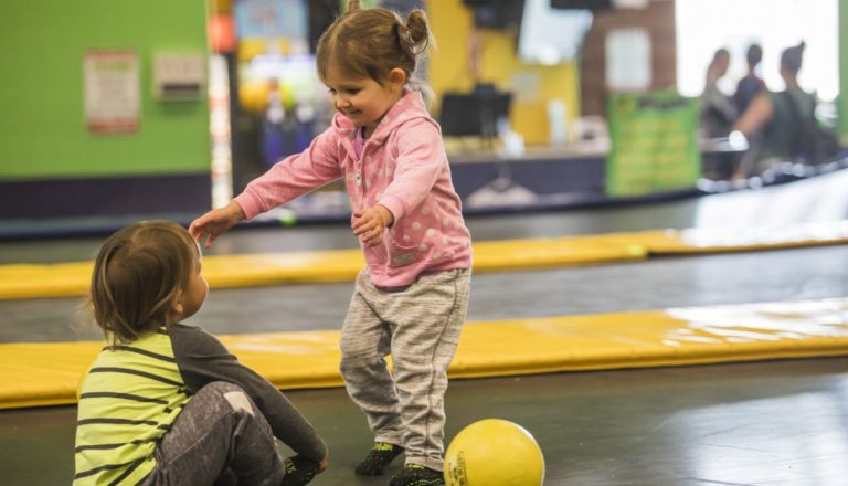 Two young children playing with a ball on trampolines.